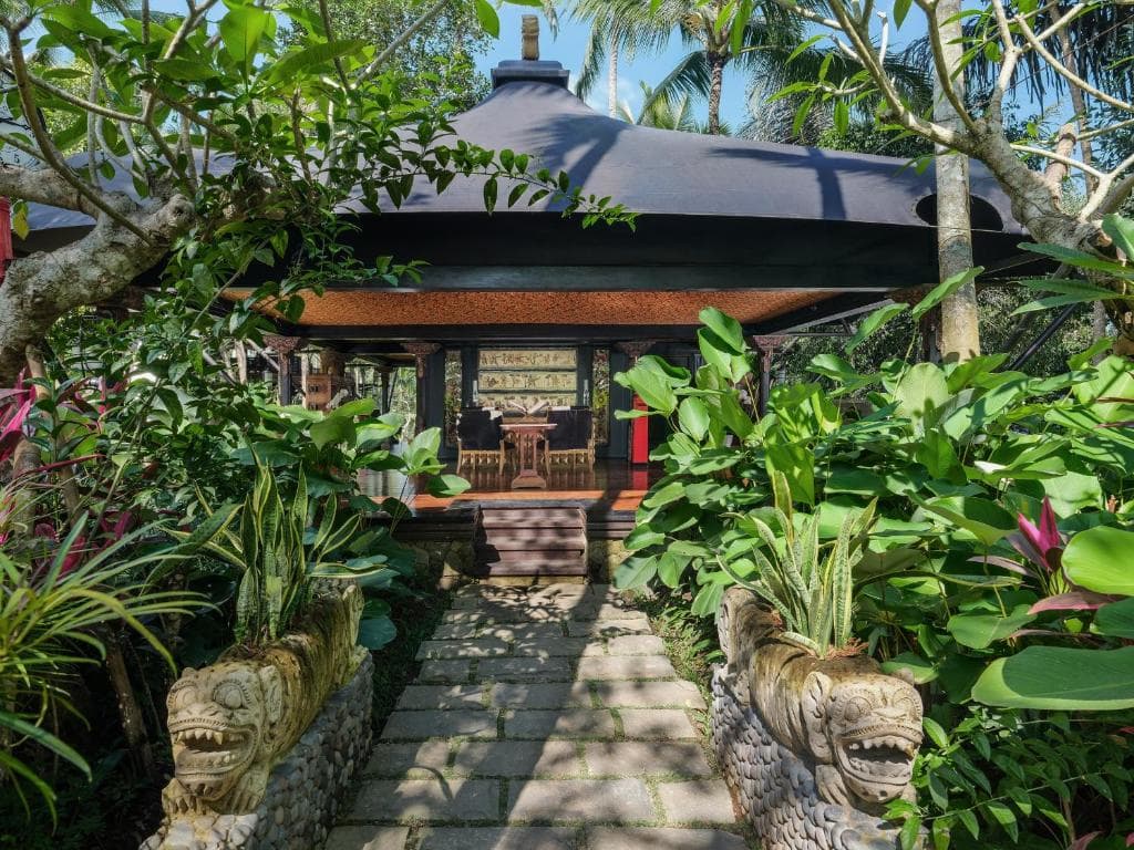 A stone pathway lined with tropical plants and carved lion statues leads to a thatched-roof pavilion with wooden doors and red accents at Capella Ubud