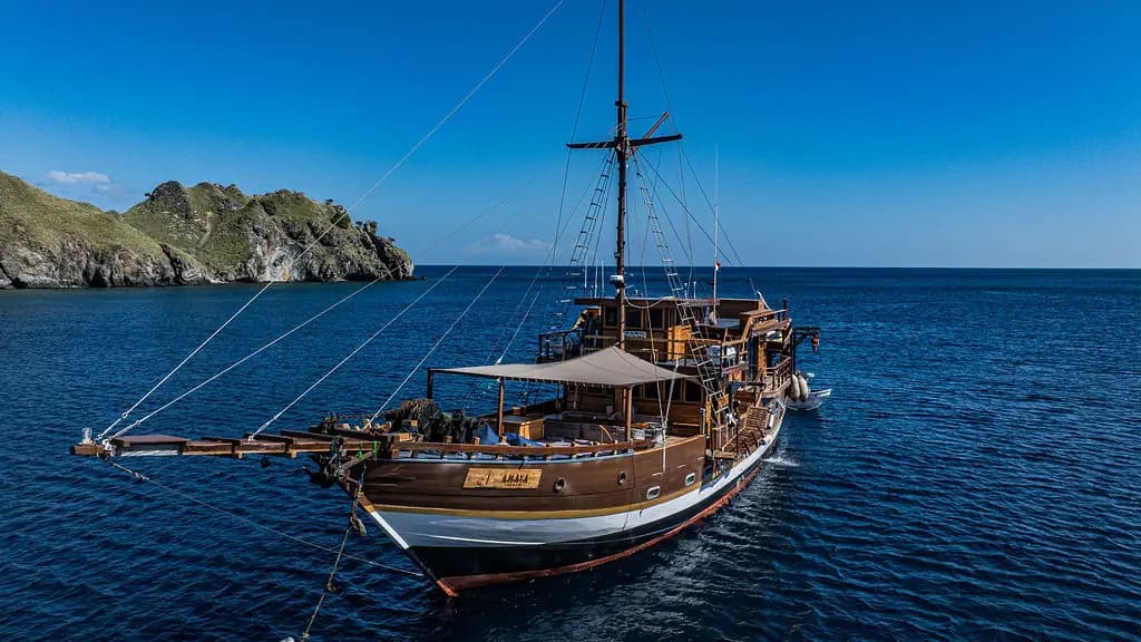 Amaya Explorer wooden liveaboard anchored near a green hilly coastline under a clear blue sky