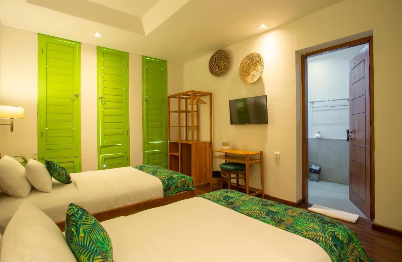 Eco Tree O'tel bedroom with two beds, green louvered doors, wooden desk, wall-mounted TV, and open door to bathroom