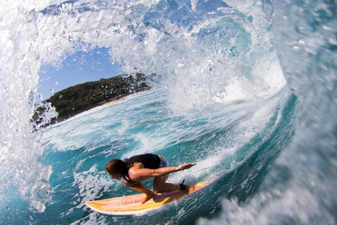 A surfer rides a wave on a yellow surfboard, captured from inside the barrel with white water and turquoise water visible, a forested headland in the distance