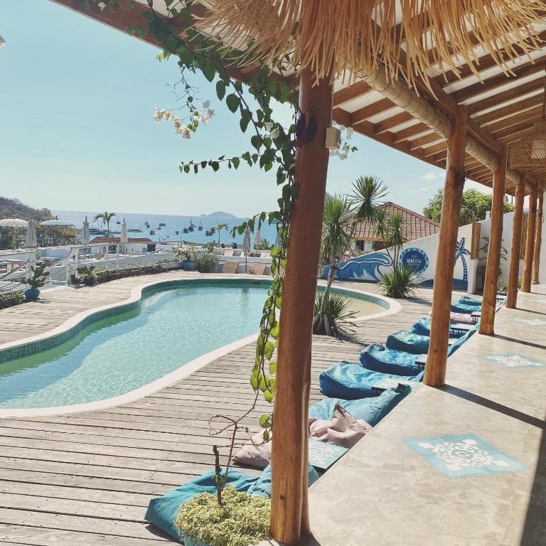 Seaesta Komodo pool area with wooden pergola, curved pool, blue seating, and a distant view outside under clear sky