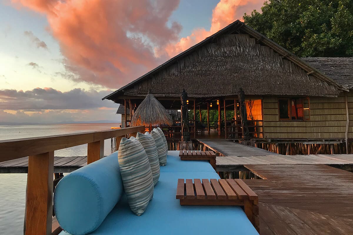 Wooden deck with blue loungers and striped pillows overlooking water, with thatched-roof bungalows at sunset