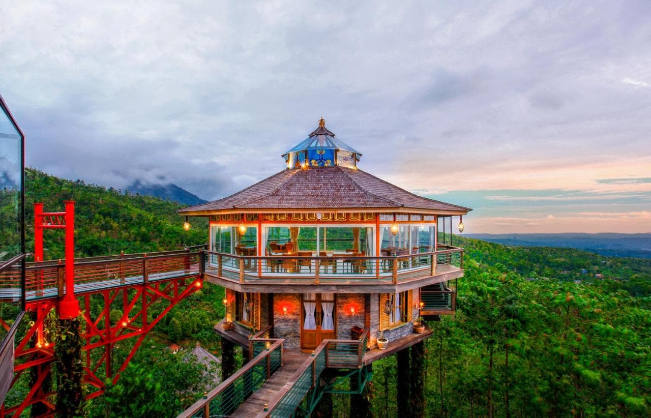 Elevated wooden pavilion with blue roof and red bridge walkway overlooking forested landscape at dusk