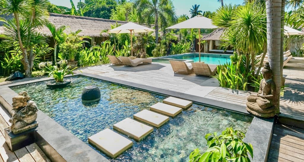 L'Archipel's pool area with stepping stones through shallow water, wooden deck, lounge seating, umbrellas, and tropical vegetation surrounding the property