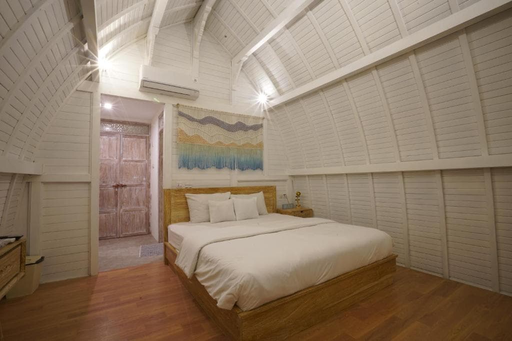 Bedroom with wooden bed, white bedding, white paneled walls and ceiling, wooden floorboards, and abstract wall art above the headboard