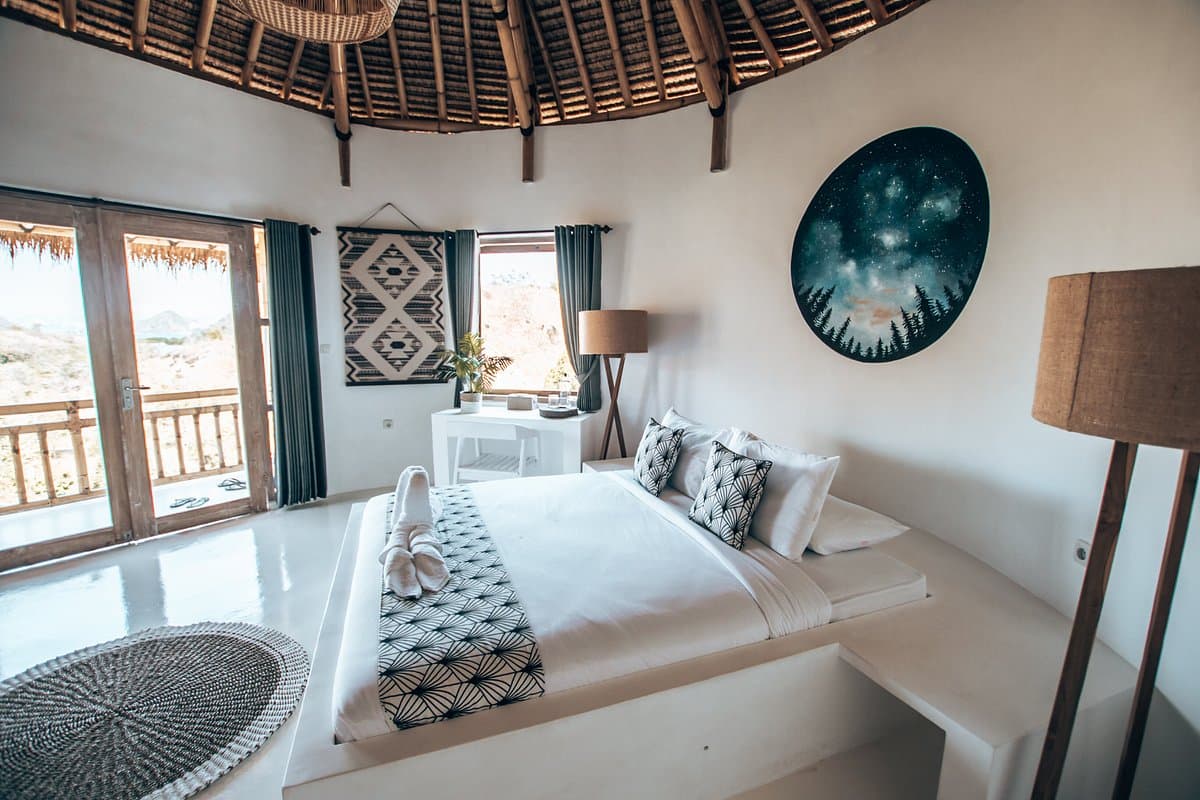 Circular bedroom with white bed, geometric patterned runner, woven ceiling, open doors to balcony, wall art, table lamp, and woven floor mat
