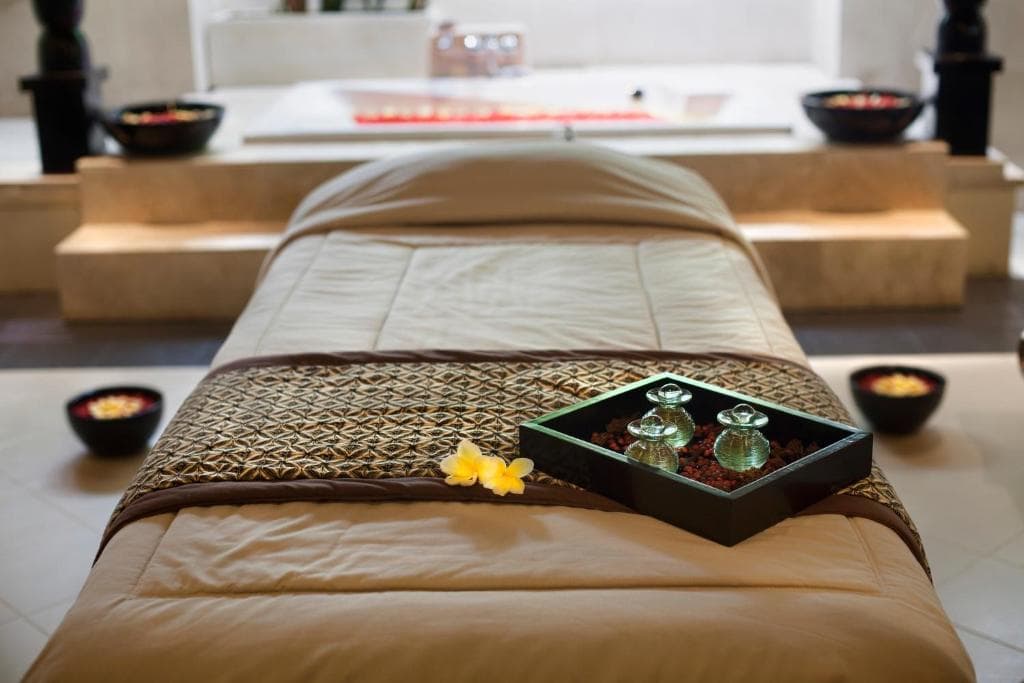 Bedroom at Puri Santrian with a low bed, woven bed runner, decorative black box with bottles, yellow flower, and small bowls on wooden shelves