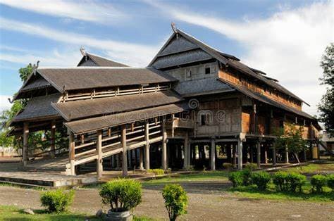 Traditional wooden stilt house with layered thatched roofs, raised on timber posts above a grassy yard