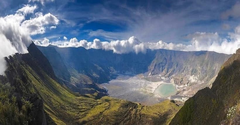 Aerial view of a volcanic caldera with a turquoise crater lake surrounded by steep green ridges and clouds