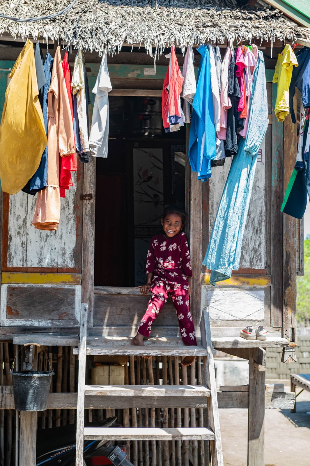 Girl smiling on a wooden stilt house porch beneath colorful laundry hanging under a thatched roof
