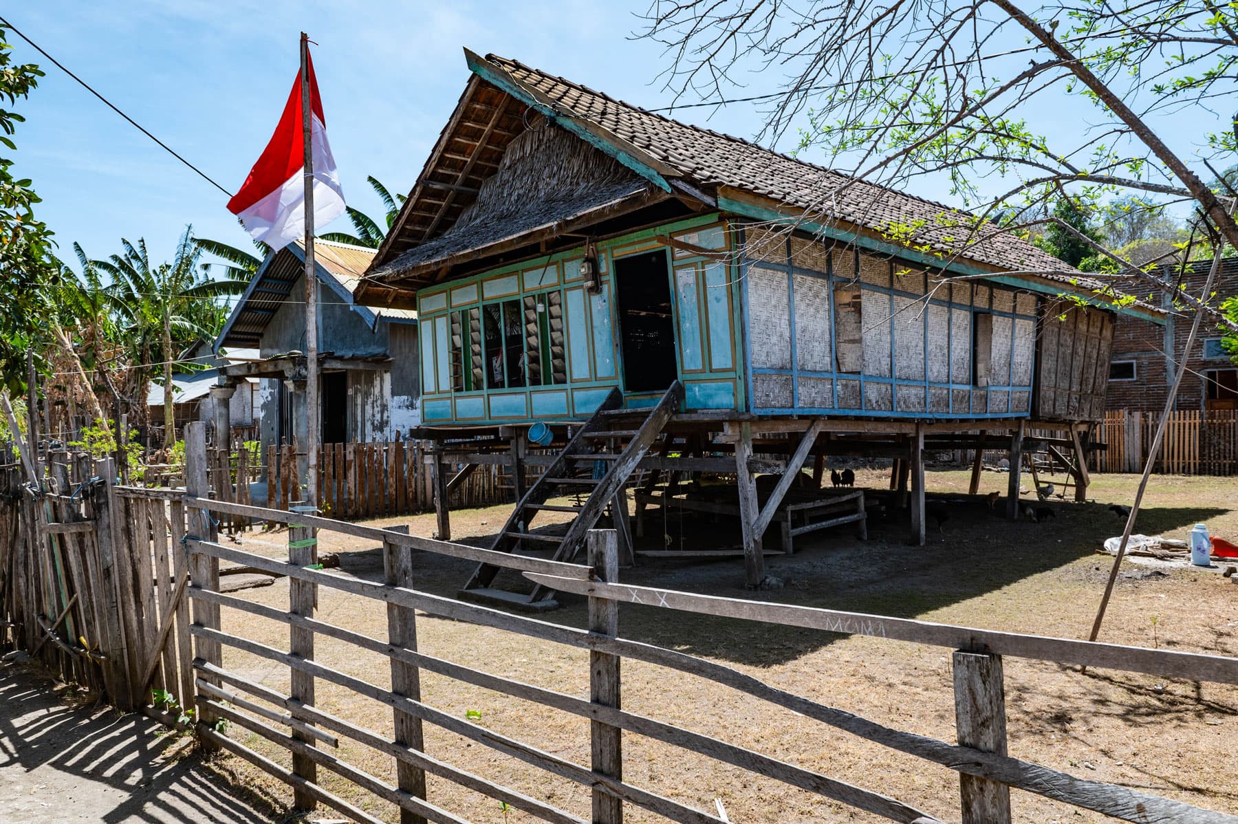 Traditional wooden stilt house with a thatched roof and Indonesian flag, enclosed by a wooden fence