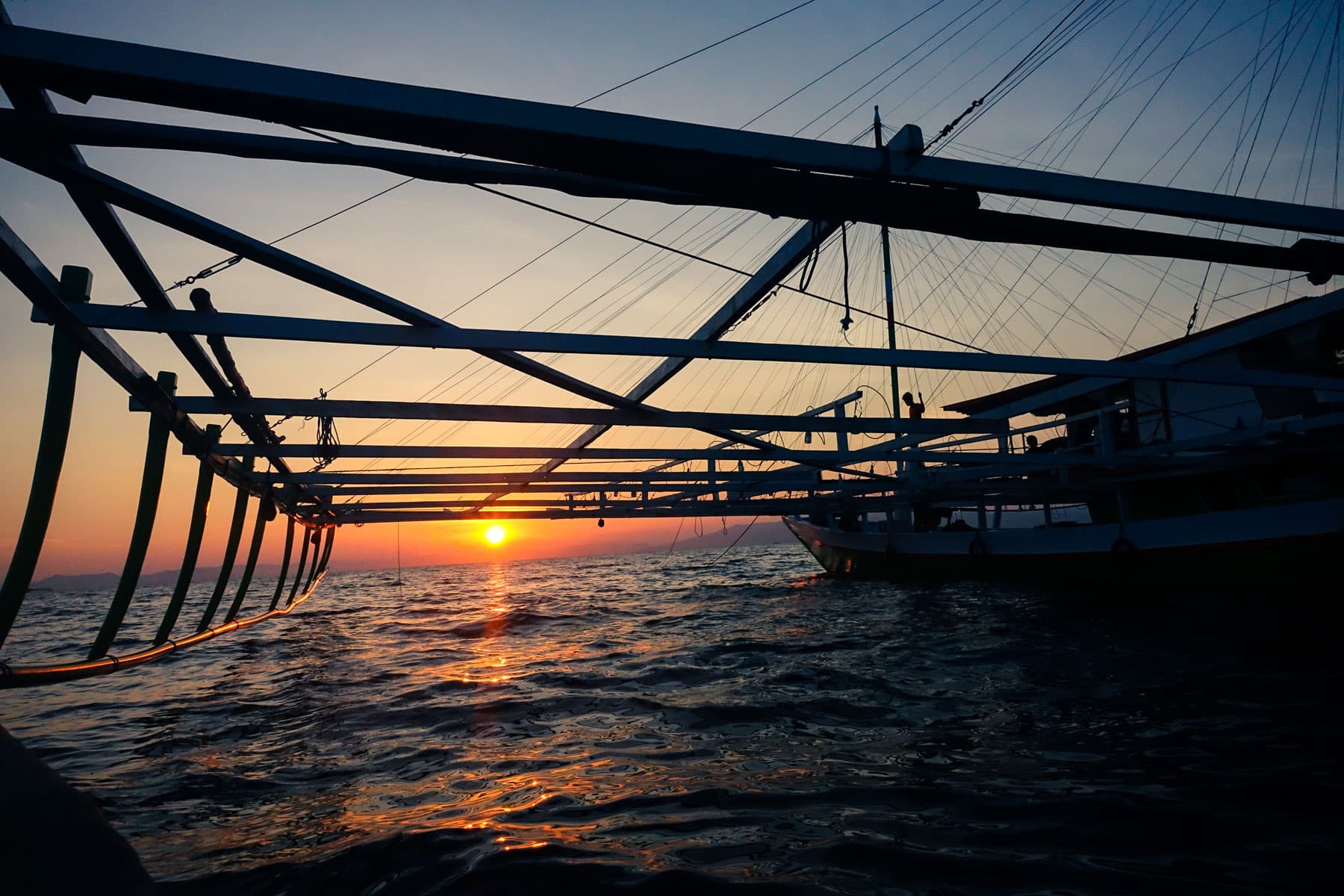 Silhouette of a traditional boat's rigging and outrigger frame against the sun setting over calm water