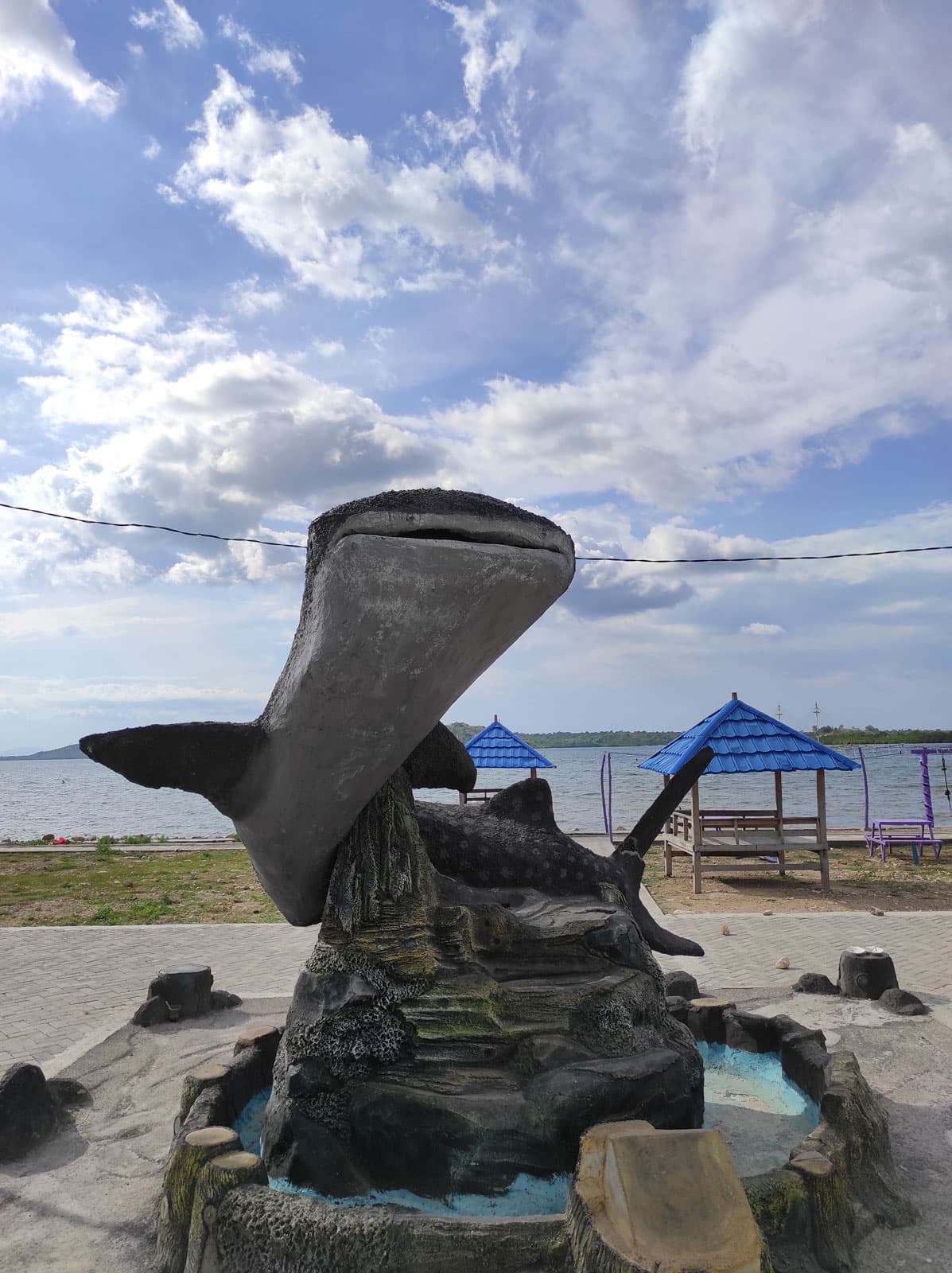 Whale shark statue on a rock fountain sculpture, with blue-roofed gazebos and the sea in the background