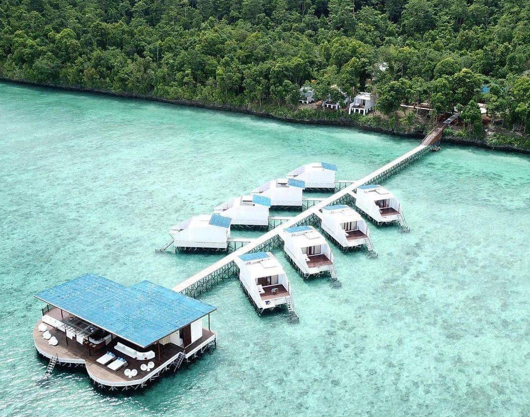 Aerial view of overwater bungalows with white roofs arranged in rows, a covered boat shelter with blue canopy, and dense forest along the shoreline