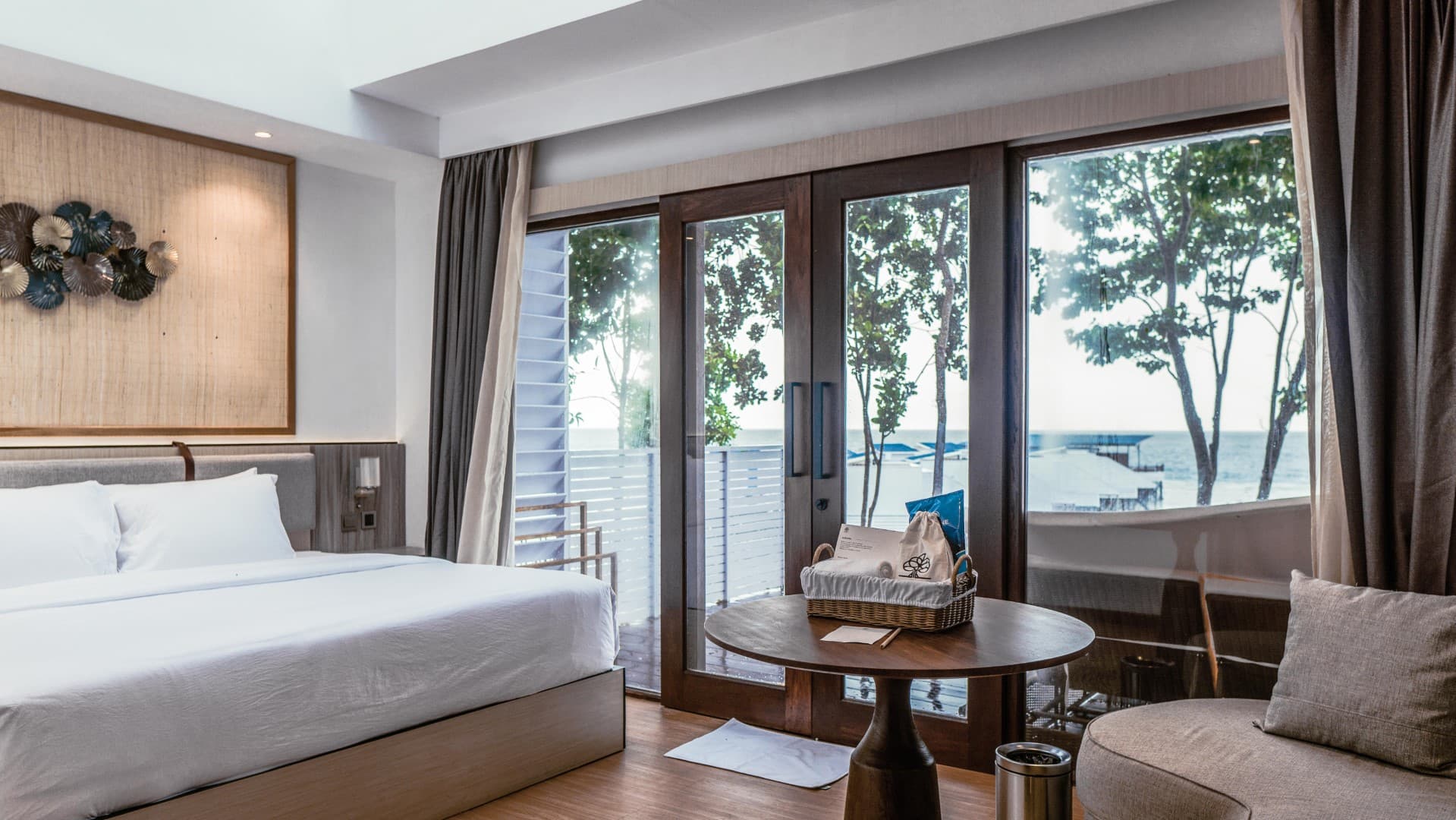 Arasatu Villas bedroom with bed, round table, seating, large windows with curtains, wooden flooring, and a view outside
