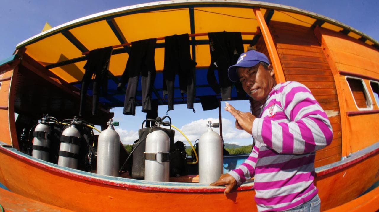 A person in a striped pink and white shirt stands beside an orange boat displaying scuba diving air tanks