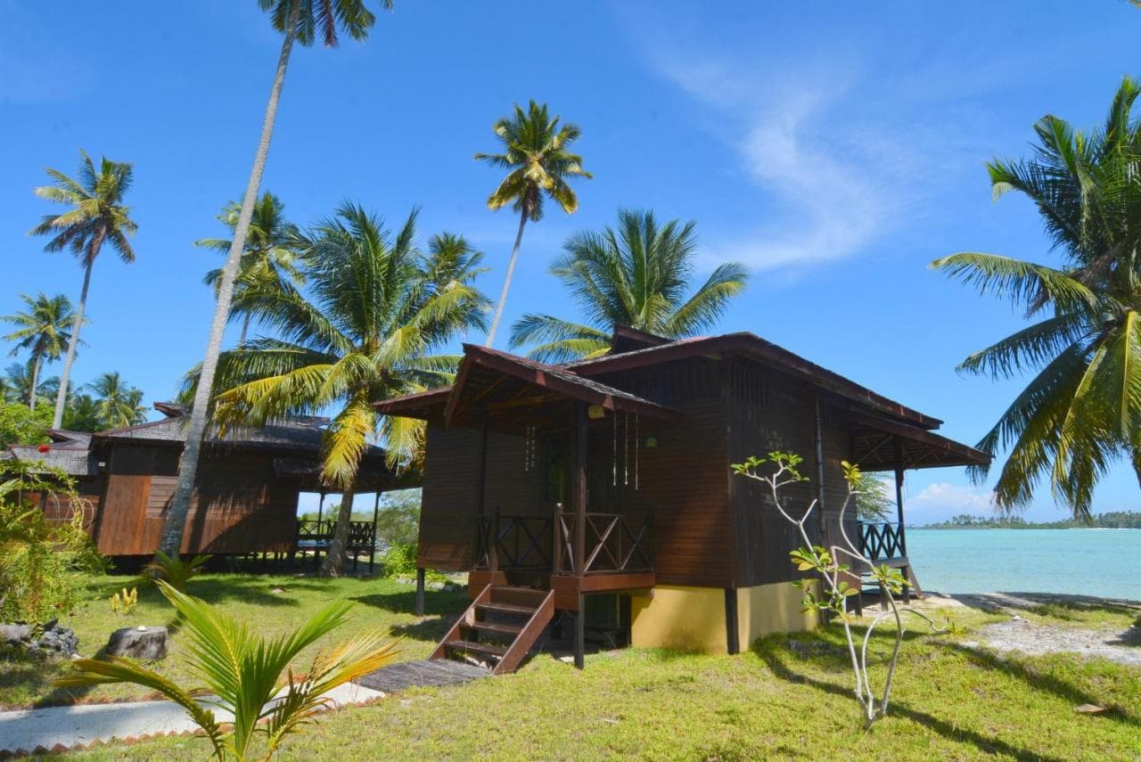 Dark wooden bungalow on stilts with a covered porch, surrounded by palm trees and green lawn, with water visible beyond