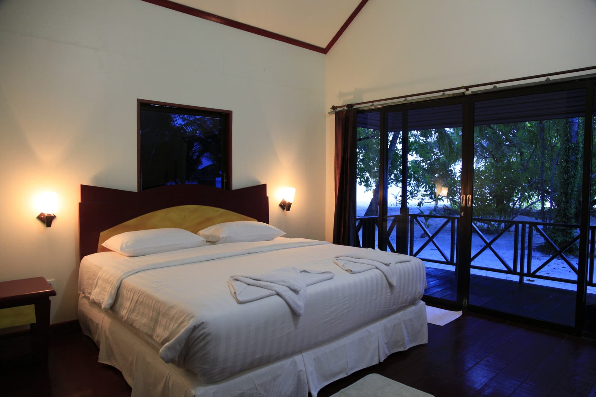 Sangalaki Resort bedroom with queen bed, white linens, bedside lamps, dark wood headboard, and glass doors opening to a balcony overlooking trees and water at dusk