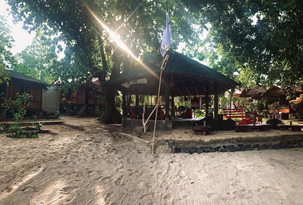 Open-air covered structures and wooden buildings with sandy grounds beneath dense tree canopy at 3Will Bunaken Dive Resort