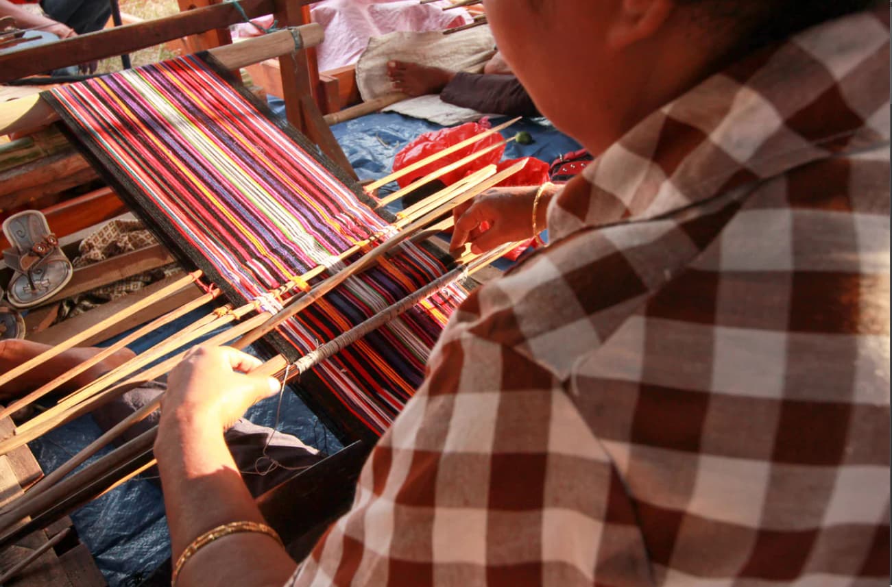 A woman weaves colorful striped fabric on a traditional handloom, wearing a checkered shirt and gold bracelets