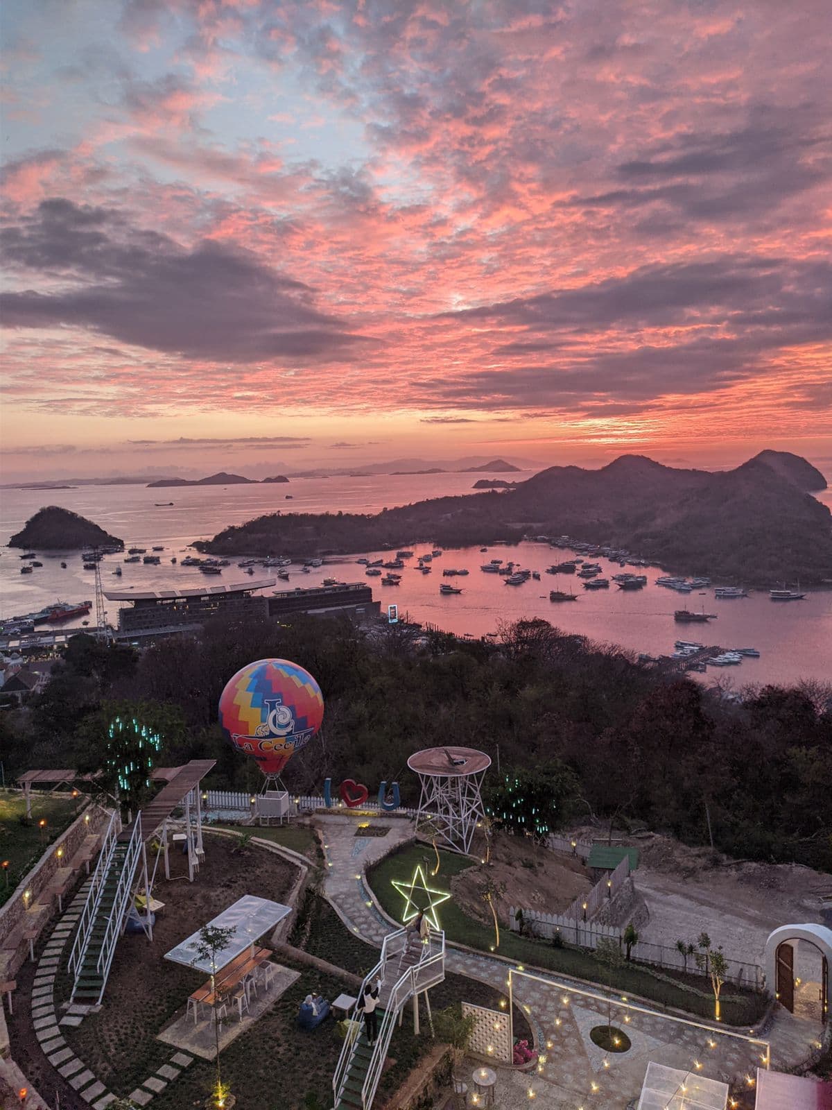 La Cecile Hotel and Cafe viewpoint at sunset with a hot air balloon, illuminated star decoration, and water with boats visible below at dusk
