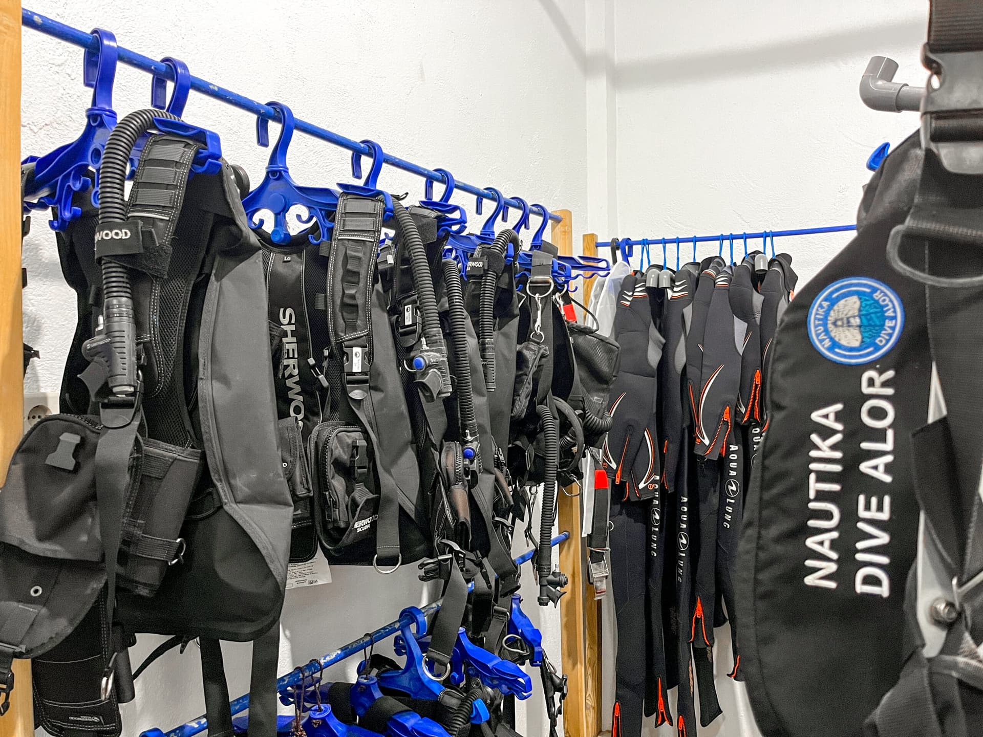 Diving equipment including multiple wetsuits and buoyancy compensators hanging on a blue rack mounted on a white wall