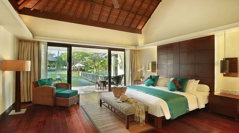 Spacious bedroom at Prama Sanur Beach Bali with twin beds, teal accents, wooden ceiling, seating area, and glass doors opening to a terrace