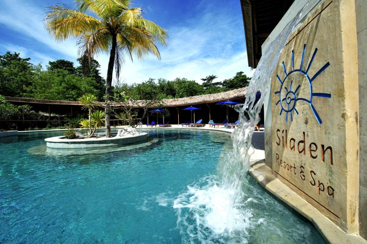 Siladen Resort and Spa's pool area with turquoise water, central island with palm tree, thatched structures, and blue umbrellas under clear sky