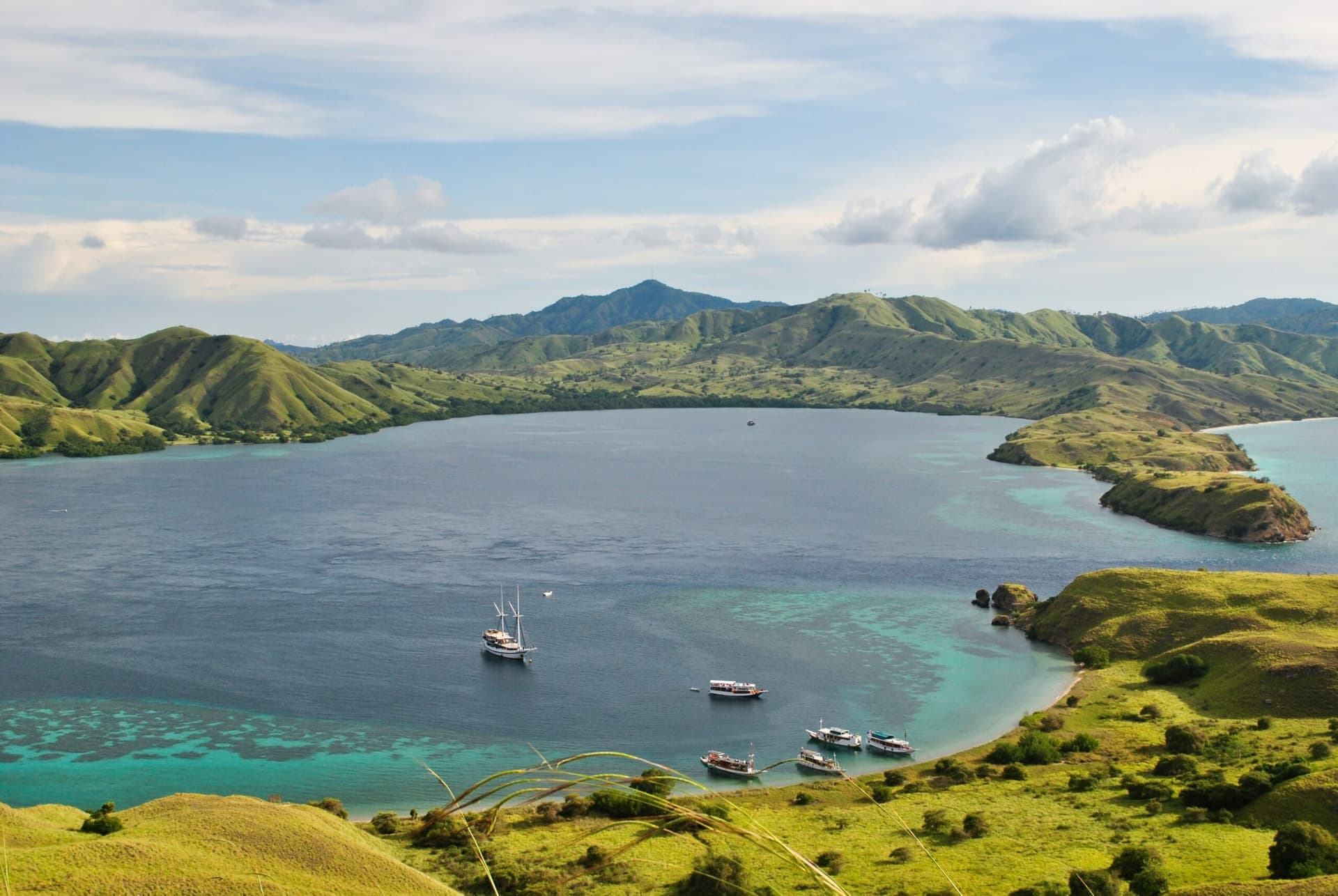 Aerial view of a sheltered bay with anchored boats, turquoise shallows, and green hills surrounding calm water