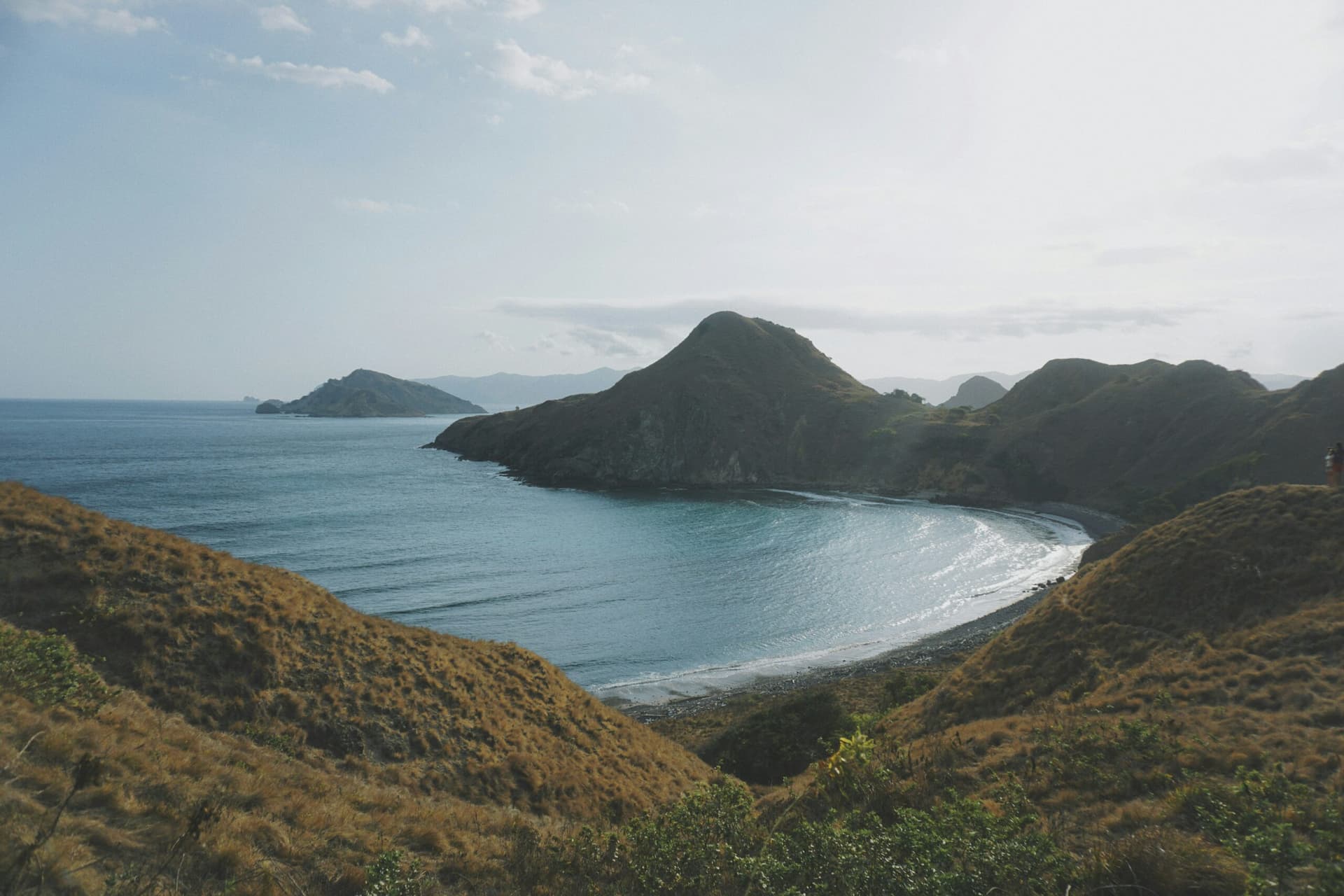 Curved bay with calm blue water surrounded by dry grassy hills and layered coastal headlands under a hazy sky