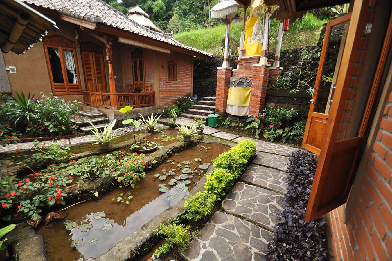 Puri Lumbung Cottages courtyard with a central pond, stone pathways, flowering plants, wooden buildings with brick details, and lush greenery surrounding the property