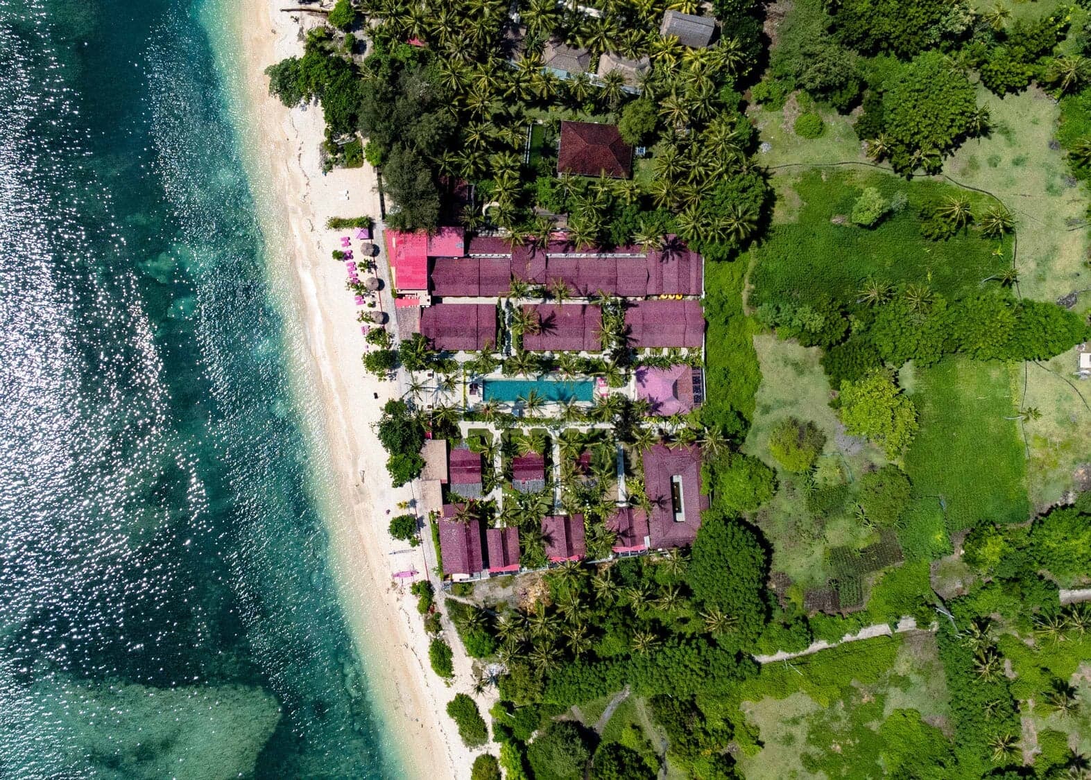 Aerial view of Pink Coco Gili Air showing pink-roofed buildings, a swimming pool, surrounding vegetation, and a sandy beach with water alongside