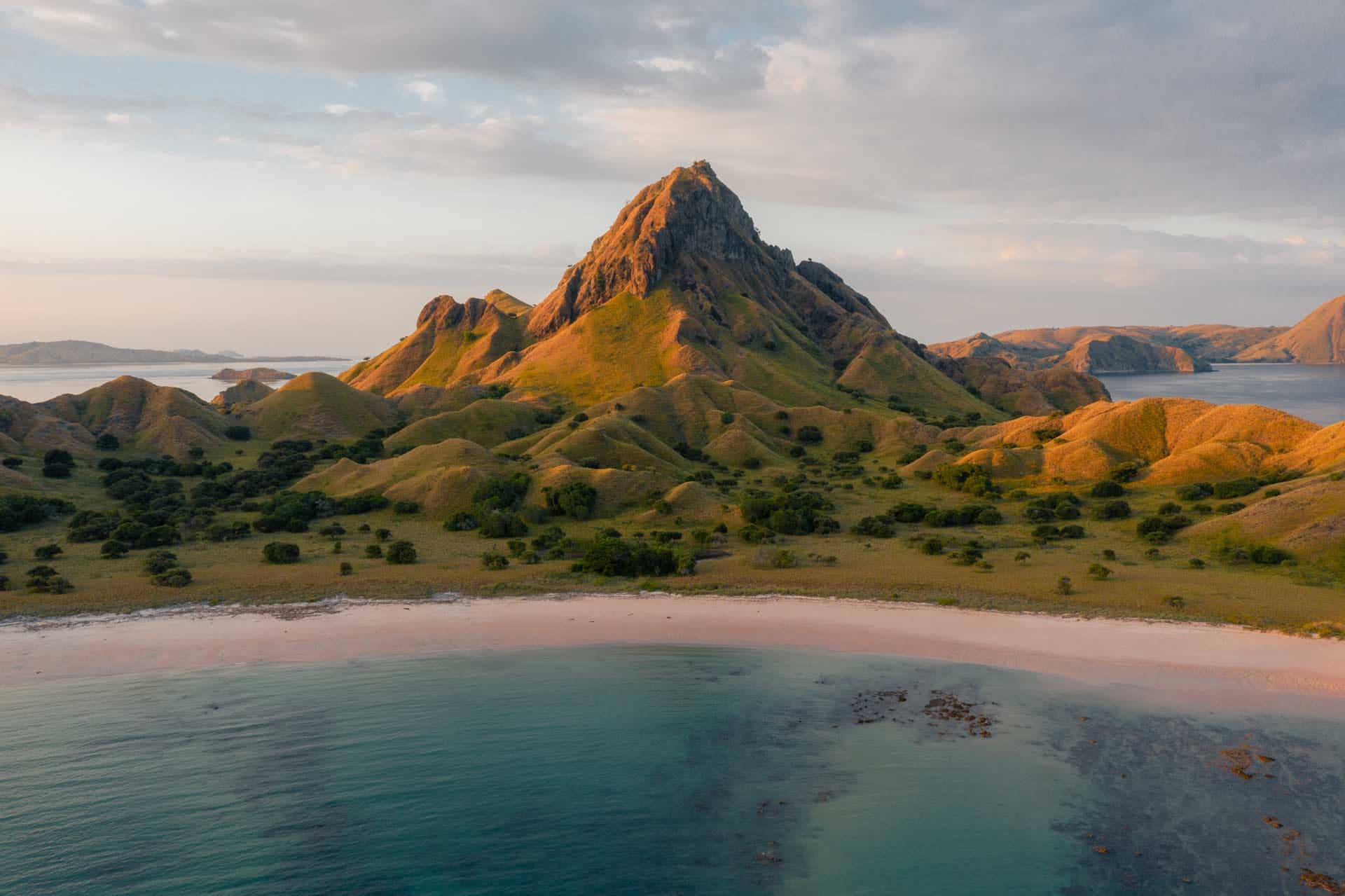 Aerial view of a jagged peak rising above green hills, with a pink-sand beach meeting turquoise water