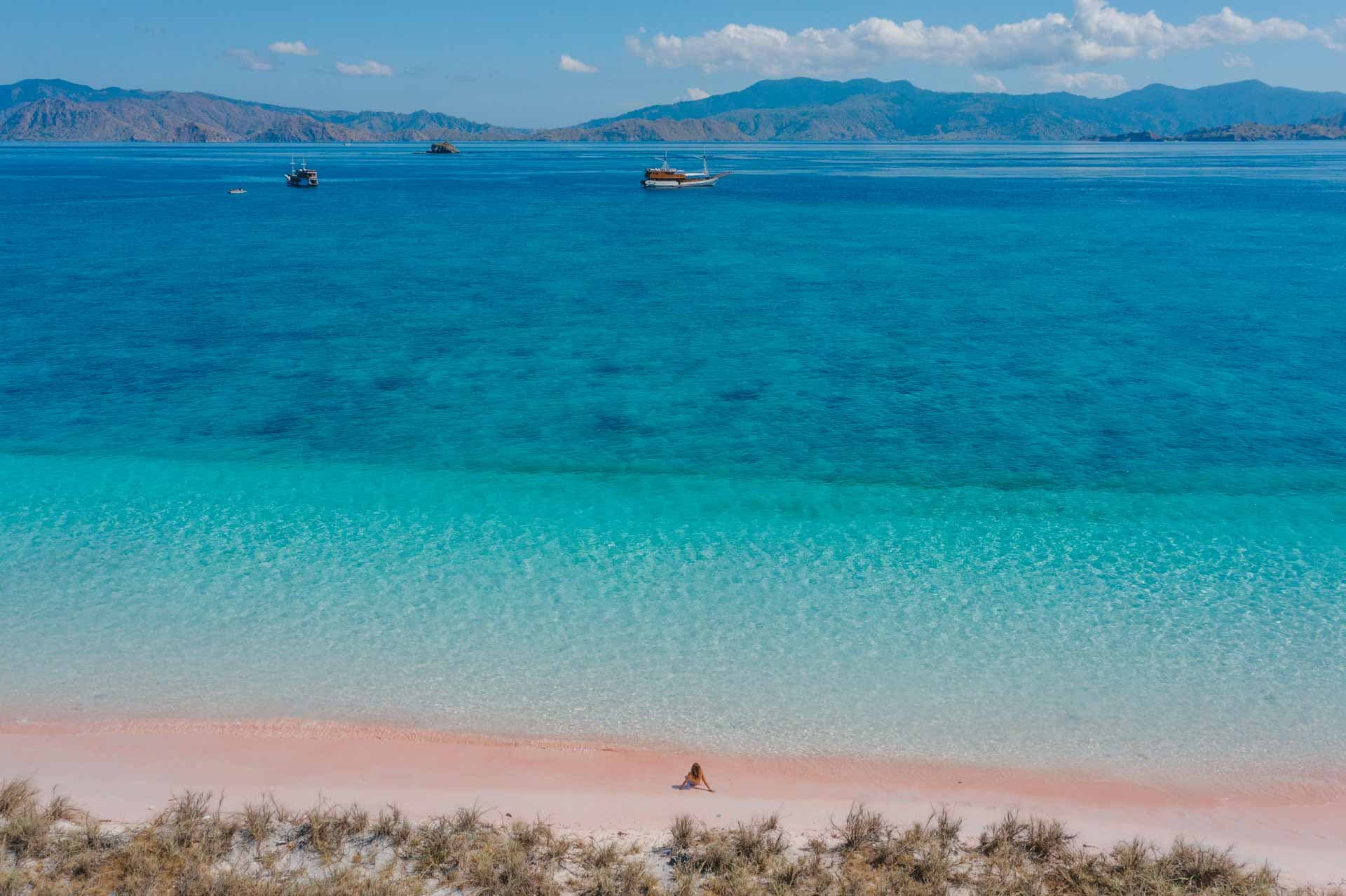 Aerial view of a pink-sand beach with turquoise water, anchored boats, and a person sitting near the shoreline