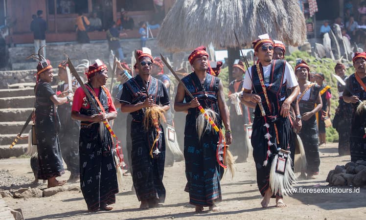 Men in traditional black woven clothing and feathered headdresses carry swords and spears during a ceremonial procession