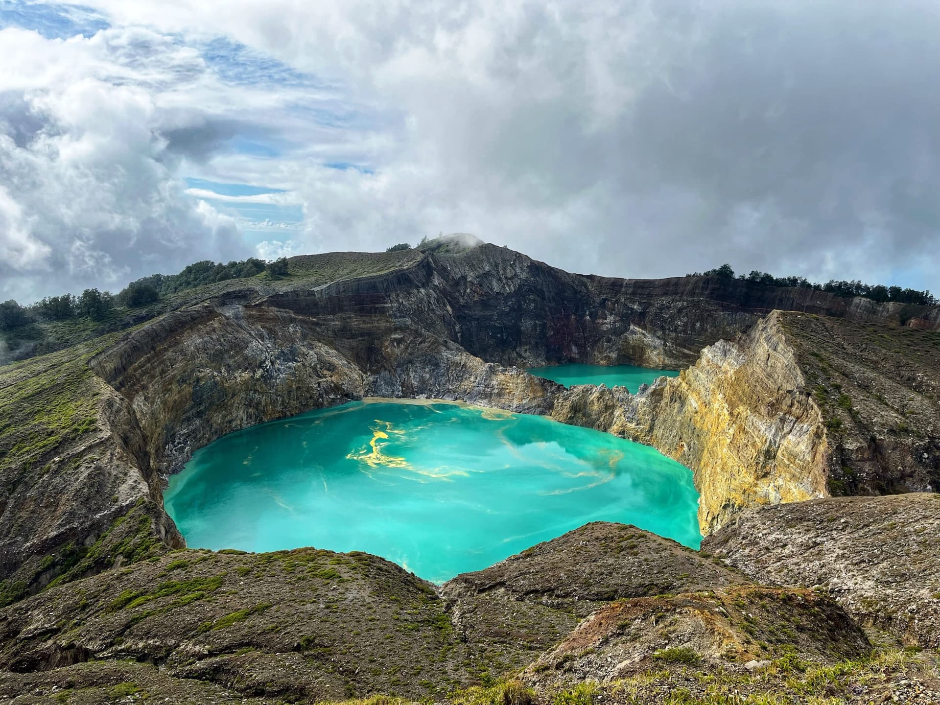 Turquoise volcanic crater lakes seen from above, surrounded by mineral-streaked cliffs under a cloudy sky