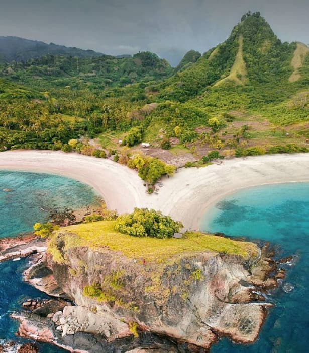 Aerial view of a rocky headland dividing two white sand beaches, with forested hills and turquoise water surrounding it