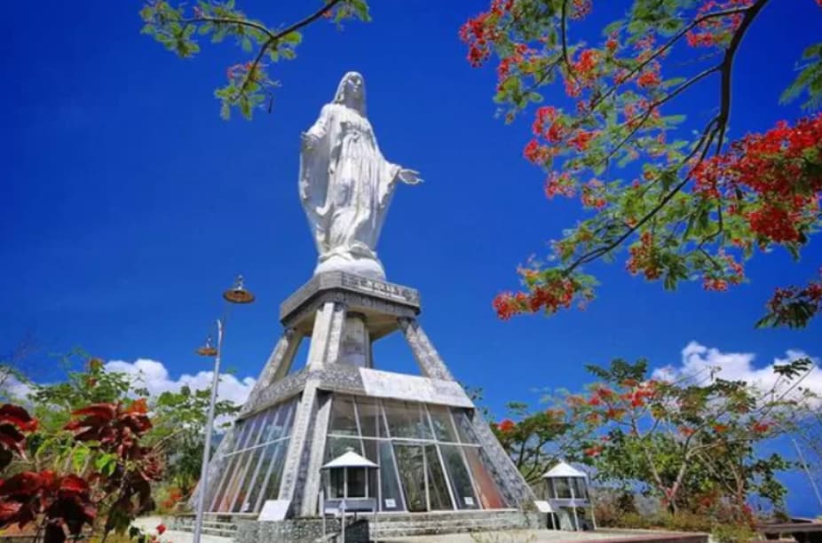 White statue of a robed figure with raised arms atop a tall triangular base, framed by red-flowered branches against a blue sky