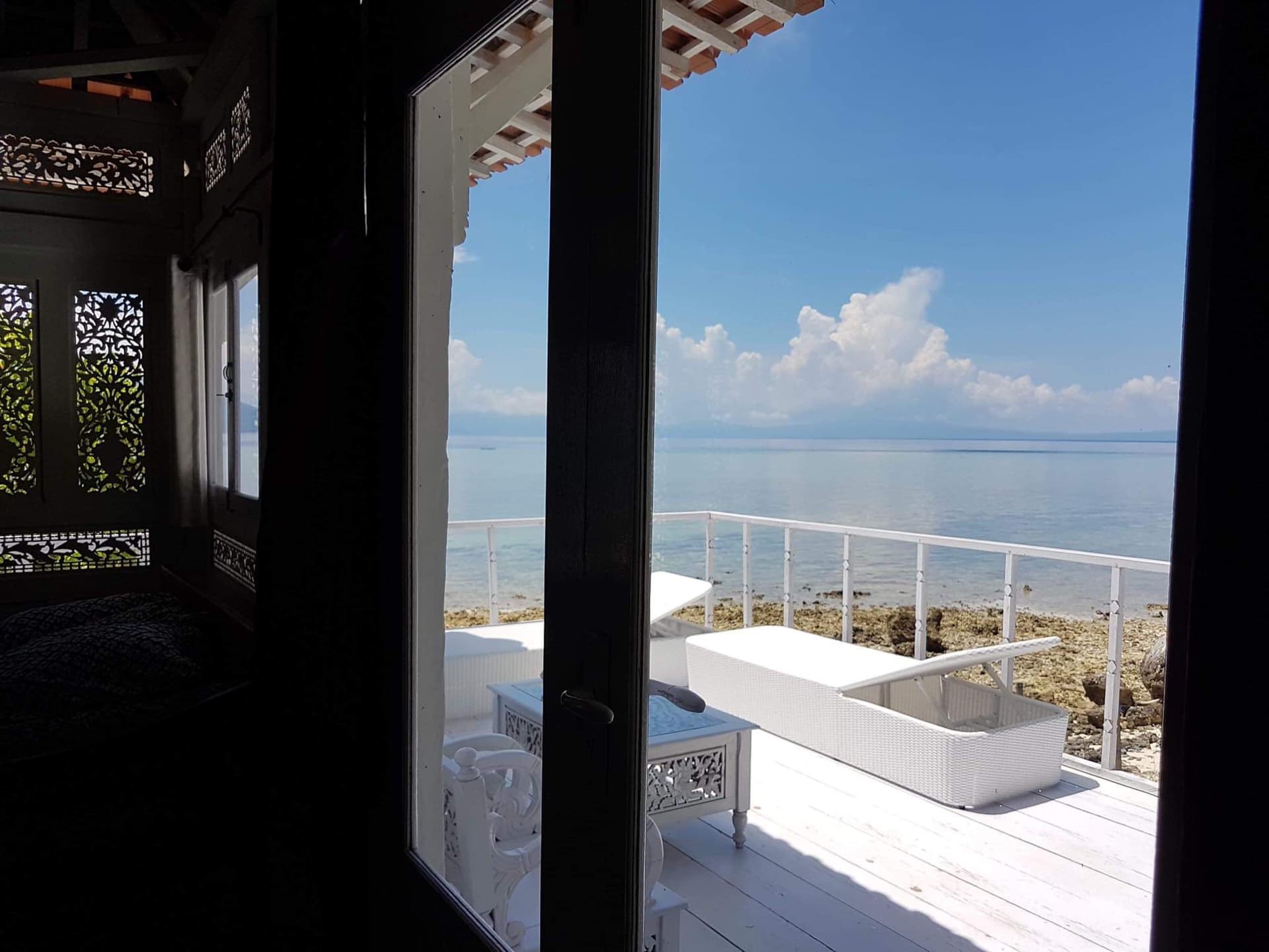 View from a darkened interior room through open glass doors to a white deck with white railings overlooking water and sky