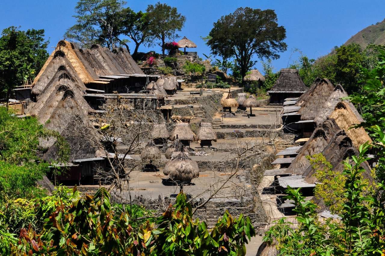Traditional thatched-roof village houses on a stepped stone hillside surrounded by trees under a clear blue sky
