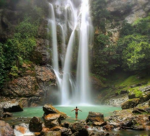 A person standing with arms outstretched in a turquoise pool beneath a tall multi-tiered waterfall surrounded by green cliffs