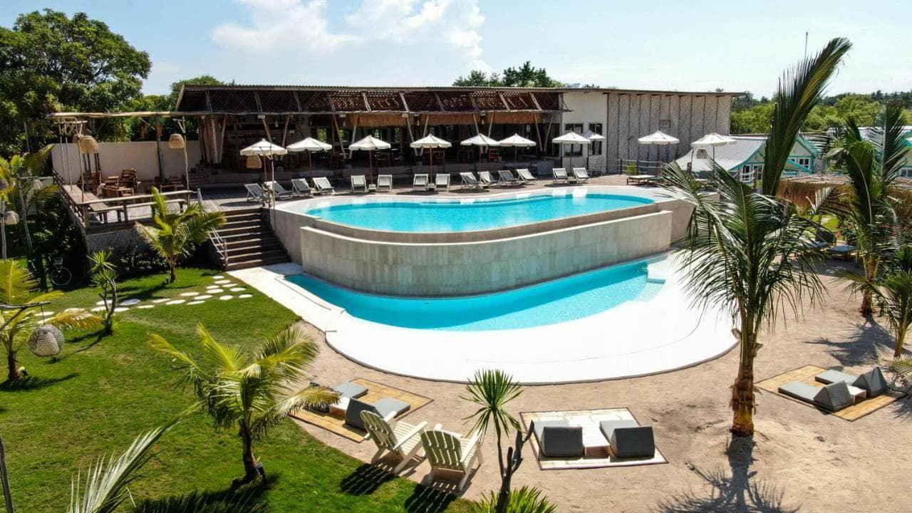 Le Pirate Gili Meno's pool area with loungers, umbrellas, manicured lawn, palm trees, and wooden structures in daylight