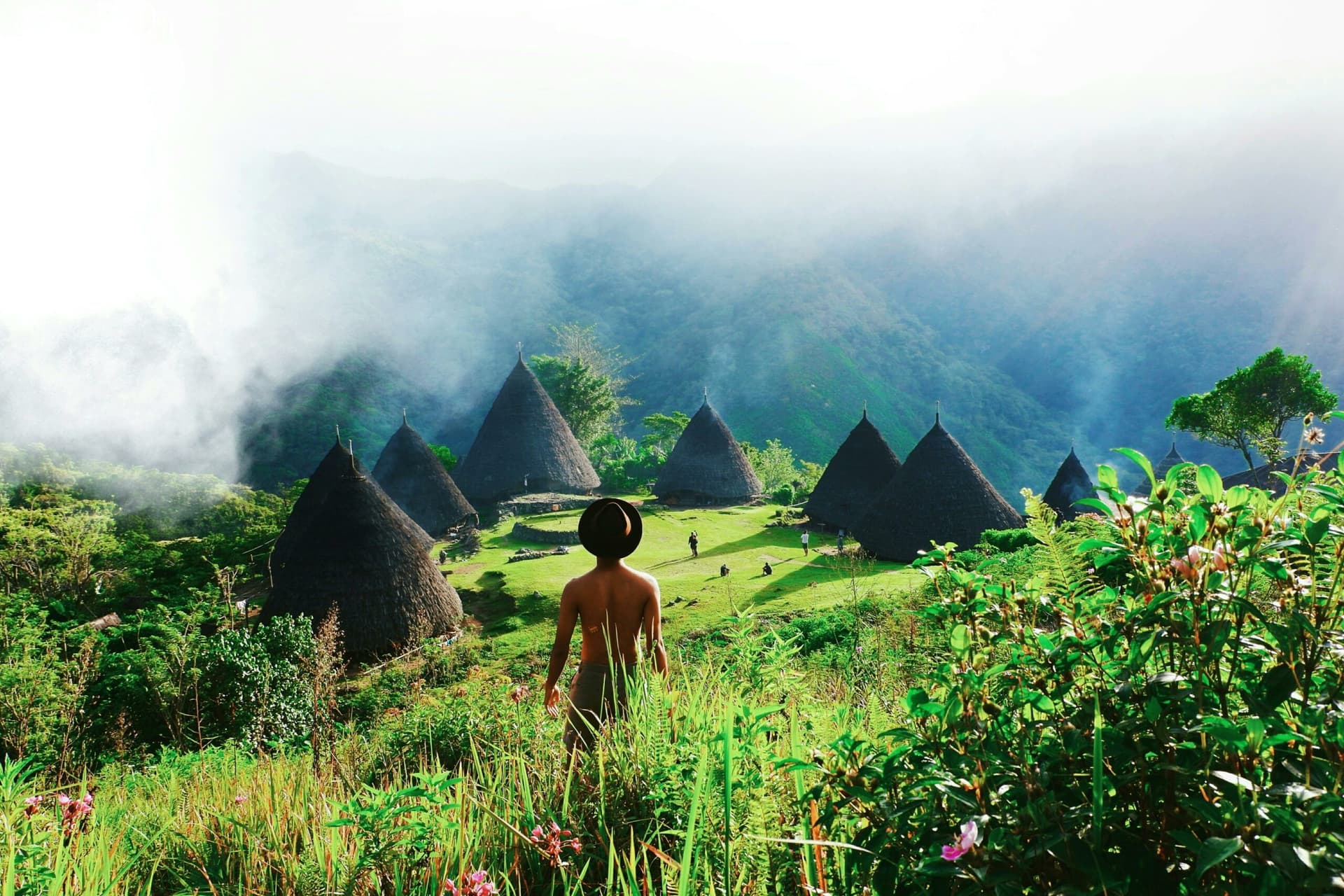 A person stands in tall grass overlooking a cluster of cone-shaped thatched-roof huts surrounded by misty green mountains
