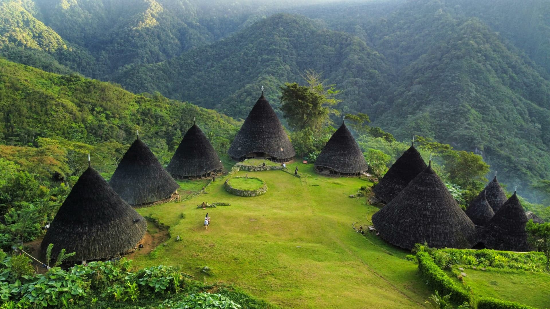 Traditional cone-shaped thatched-roof huts arranged around a grassy clearing surrounded by dense forested mountains