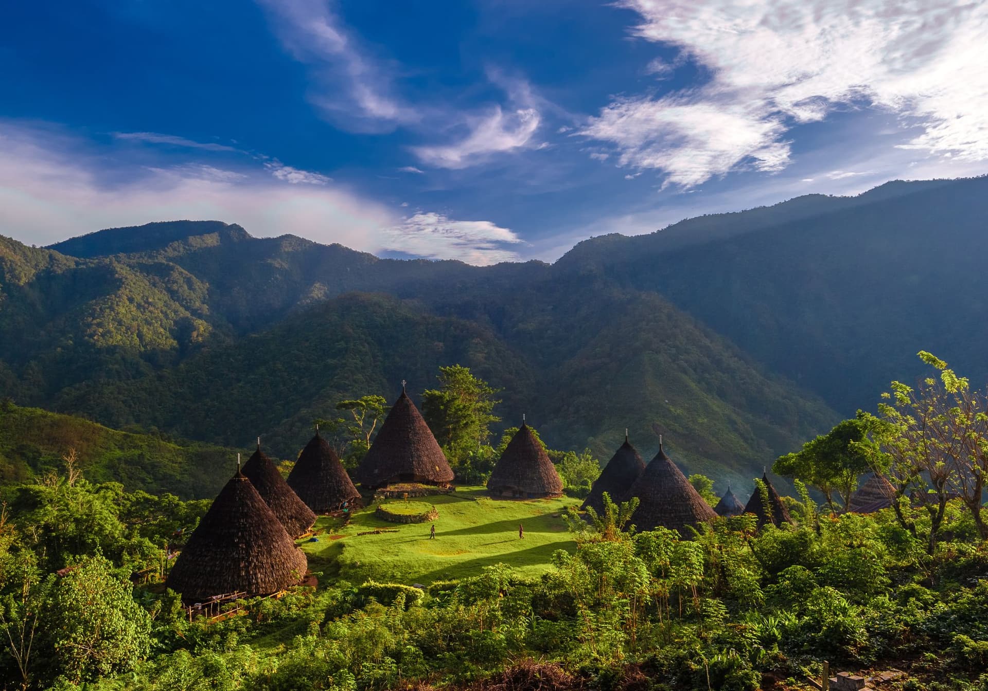 Traditional cone-shaped thatched-roof huts on a grassy clearing surrounded by forested mountains under a blue sky