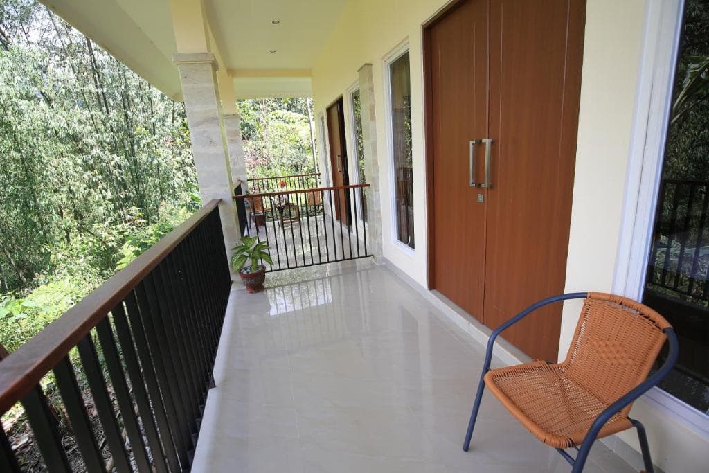 Covered veranda with white flooring, brown wooden door, orange chair, potted plant, black railing, and green vegetation visible beyond