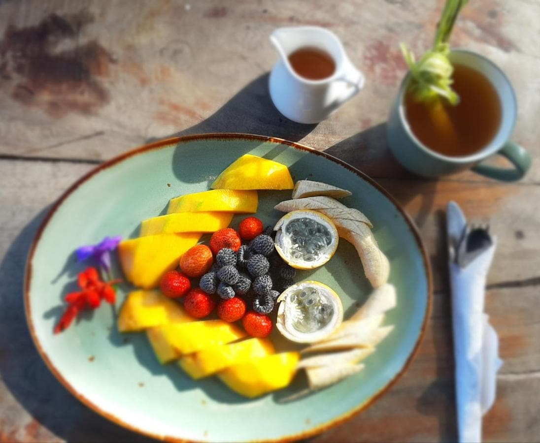 Breakfast plate at Gumi Ayu Ecostay with sliced mango, berries, lemon halves, edible flowers, and pastries, served with tea and honey