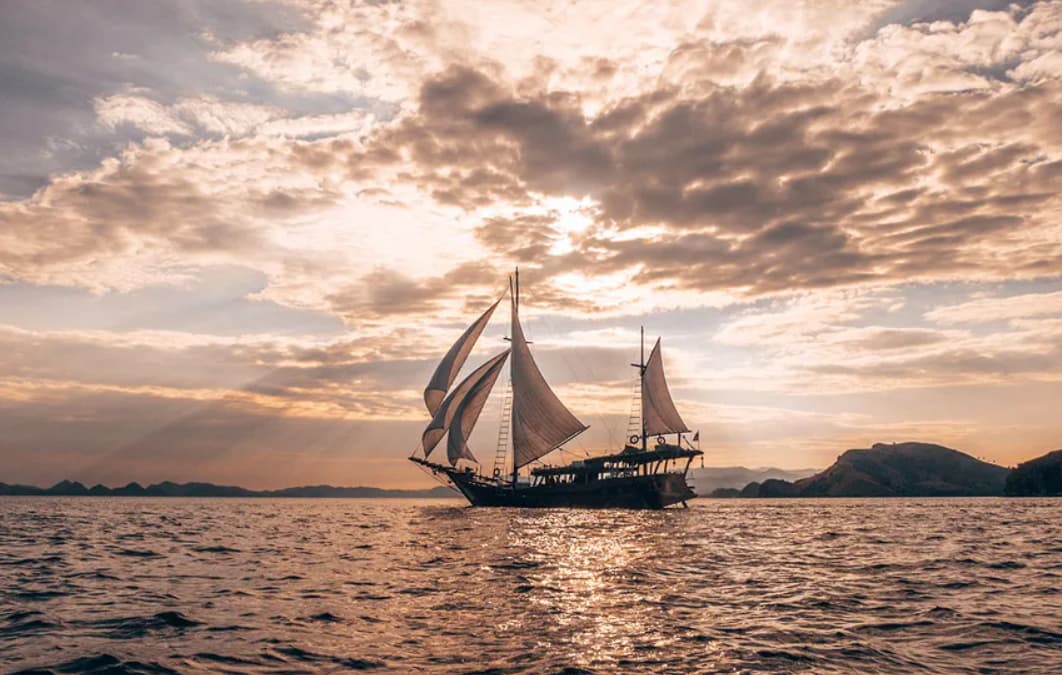 Nataraja liveaboard under full sail on calm water at sunset, silhouetted against a cloudy sky with distant hills