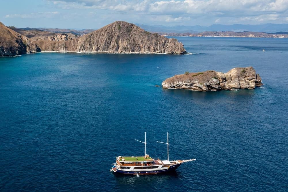 Aerial view of the Indo Master liveaboard anchored near a small rocky island with hills in the background