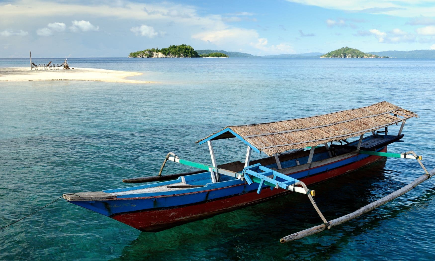 Traditional outrigger boat with a thatched roof anchored in clear water near a small forested island